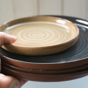 May include: A stack of three ceramic plates featuring a small, light tan plate with a spiral pattern on top, resting on two larger, dark charcoal-colored plates with a smooth, glossy finish and visible terracotta-colored clay edges.