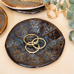 May include: Three gold rings with a green gemstone on a dark blue ceramic ring dish with a geometric pattern.