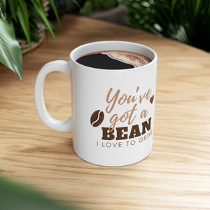May include: White ceramic coffee mug filled with dark coffee. The mug has a curved handle and features the text "You've got a BEAN I LOVE TO GRIND" in brown and tan lettering, along with a coffee bean graphic.