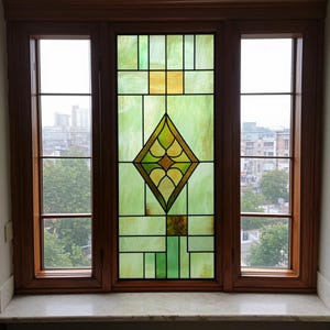 Puede incluir: Una ventana de tres paneles con un panel central de vidrieras. La vidriera presenta un diseño de diamante con detalles amarillos y verdes, rodeado de formas geométricas verdes y amarillas. El marco de la ventana es de madera oscura.