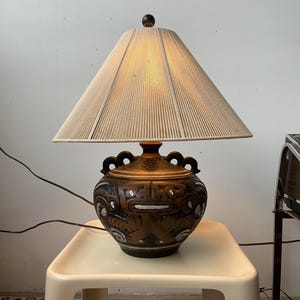 Peut inclure: Lampe de table décorative avec un socle en céramique marron orné de motifs sculptés et un abat-jour beige. La lampe est posée sur une petite table blanc cassé. Le luminaire a un fleuron marron foncé.