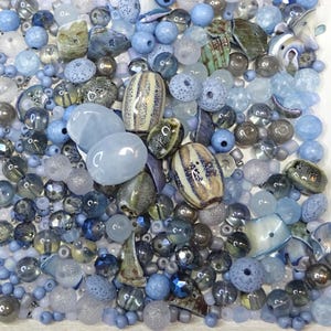 Könnte beinhalten: Eine Ansammlung blauer Perlen und dekorativer Elemente in verschiedenen Formen und Größen. Die Kollektion umfasst runde, ovale und unregelmäßige Perlen in den Farbtönen Hellblau, Marineblau und Grau, mit strukturierten und gemusterten Oberflächen.