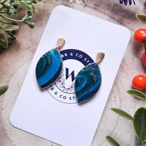 May include: A pair of teardrop-shaped earrings with a gold-colored top and a marbled blue and teal design. The earrings are displayed on a white card with a logo that reads "BB & CO ST". The background includes greenery and berries.