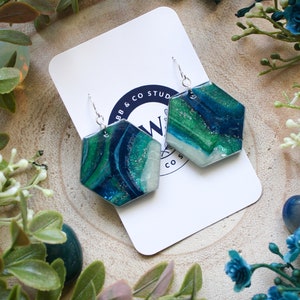 May include: A pair of silver earrings with hexagonal green, blue and white resin pendants. The resin has a marbled effect and glitter accents.