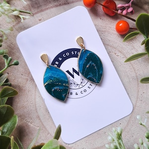 May include: Leaf-shaped earrings with a gold-coloured top and a marbled teal and gold design. The earrings are displayed on a white card with a circular logo. The background includes greenery and berries.