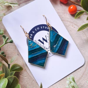 Könnte beinhalten: Ein Paar geometrische Ohrringe mit goldfarbenem Rahmen und einem marmorierten türkisblauen und blauen Design. Die Ohrringe hängen an einer weißen Karte mit dem Text "BB & CO STUD" und einem stilisierten "W"-Logo. Der Hintergrund umfasst dekoratives Grün und Beeren.