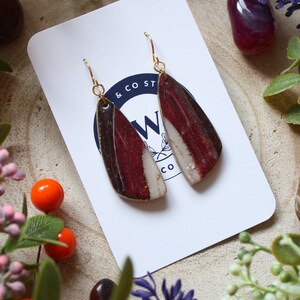 May include: A pair of dangle earrings with a white, red, and brown abstract design. The earrings are made of ceramic and have gold-toned hooks.