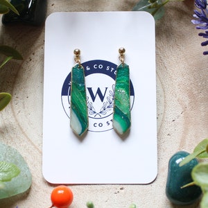 May include: A pair of green and white agate earrings with gold hooks. The earrings are shaped like long, thin teardrops.