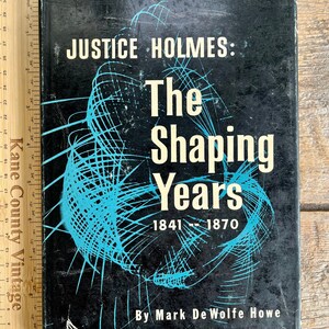 May include: A vintage book titled "JUSTICE HOLMES: The Shaping Years 1841-1870" with a black cover and white and blue text. The cover features a blue abstract design. The book is placed on a wooden surface.