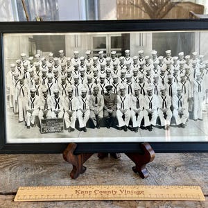 Puede incluir: Fotografía en blanco y negro de un grupo de hombres con uniforme militar. Los hombres están de pie en filas y todos llevan sombreros blancos y camisas blancas con corbatas negras. La foto está enmarcada en un marco negro y está sobre un caballete de madera.
