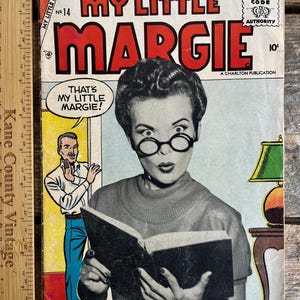 Może przedstawiać: Vintage okładka komiksu "My Little Margie" nr 14. Okładka przedstawia czarno-biały obraz kobiety w okularach czytającej książkę, z zaskoczoną miną. Tytuł jest dużymi czerwonymi literami.