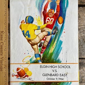 Könnte beinhalten: Ein Vintage-Programm für ein Footballspiel zwischen der Elgin High School und Glenbard East. Das Programm zeigt eine farbenfrohe Illustration von zwei Footballspielern in Aktion. Der Text auf dem Programm lautet "Elgin High School vs. Glenbard East October 9, 1964".