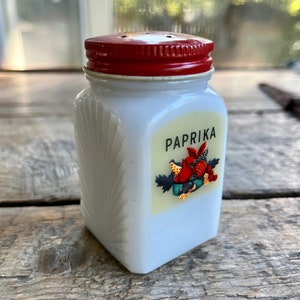 Op de afbeelding: Witte glazen strooier met een rood deksel en een etiket met de tekst "Paprika". Het etiket heeft een afbeelding van rode paprika's en andere specerijen.