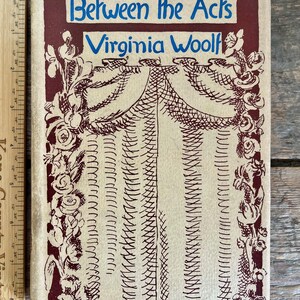 Puede incluir: Una cubierta de libro vintage en colores rojo y crema. El título "Between the Acts" de Virginia Woolf está impreso en letras azules. La cubierta presenta una ilustración estilizada de un telón de escenario con un borde decorativo.