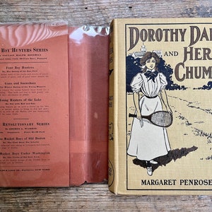 Peut inclure: Une couverture de livre vintage avec un fond jaune et du texte noir. Le titre est "Dorothy Dale and Her Chums" par Margaret Penrose. La couverture présente une illustration en noir et blanc d'une fille en robe blanche tenant une raquette de tennis.