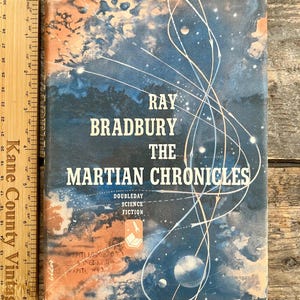 Pode incluir: Livro antigo, "The Martian Chronicles" de Ray Bradbury. A capa apresenta uma ilustração espacial azul e laranja com linhas brancas e texto. Publicado por Doubleday, ficção científica.