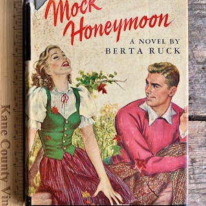 May include: A vintage paperback book cover with a red title "Mock Honeymoon" and the author's name "Berta Ruck". The cover features a couple in a forest setting with fall foliage. The woman is wearing a green vest and red and white striped skirt. The man is wearing a pink sweater.