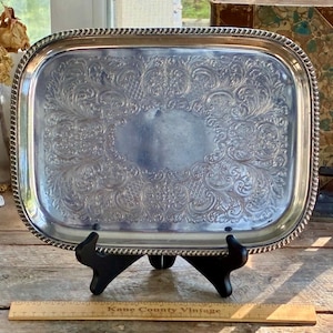 May include: A rectangular silver-toned metal tray featuring an ornate embossed floral and scrollwork pattern along the border and a smooth, plain center. The tray has a rope-style edge and is displayed on a black stand with a wooden ruler below.