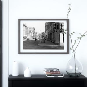 May include: Black and white photograph of a city street scene with buildings on both sides of the street. The street is empty except for a few cars and a person walking on the sidewalk.