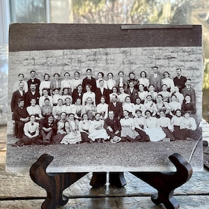 Może przedstawiać: Zdjecie w stylu vintage, czarno-białe, przedstawiające dużą grupę ludzi, prawdopodobnie z końca XIX lub początku XX wieku. Osoby są formalnie ubrane i pozują przed kamiennym murem. Zdjęcie jest prezentowane na drewnianym sztaludze.