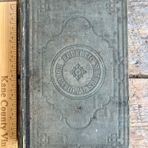 Może przedstawiać: Antyczna, zniszczona książka z wyblakłą, niebiesko-zieloną okładką. Okładka posiada wytłoczone wzory dekoracyjne i napis "ECLECTIC EDUCATIONAL SERIES" w okrągłym wzorze. Obok książki umieszczono drewnianą linijkę.