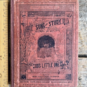 Puede incluir: Un libro rojo titulado "Song and Study for God's Little Ones" con una ilustración en blanco y negro de un niño en la portada. El libro fue publicado por R. R. McCabe & Co. en Chicago.