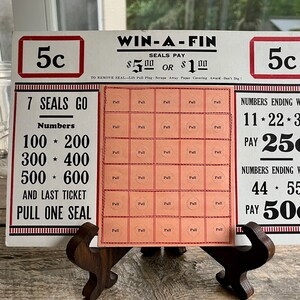 Può includere: Una carta da gioco vintage Win-A-Fin con un bordo rosso e bianco. La carta ha una griglia di 35 linguette da tirare con uno sfondo rosso e linguette da tirare bianche. La carta ha anche testo che dice "Win-A-Fin", "Seals Pay 5,00 € o 1,00 €", "7 Seals Go", "Numbers 100 * 200, 300 * 400, 500 * 600", "And Last Ticket Pull One Seal", "Numbers Ending With 11 * 22 * 33 Pay 25c", "Numbers Ending With 44 * 55 Pay 50c".