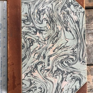 May include: A vintage book with a marbled cover in shades of green, black, and pink. The book has a brown leather spine and corners.