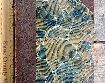 May include: Antique book with a brown leather spine and corners. The cover features a marbled design in shades of blue, green, and tan. The book measures approximately 9 inches in height and 6 inches in width.