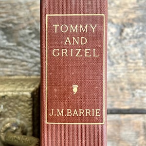Peut inclure: Un livre rouge avec des lettres dorées sur le dos. Le titre est "Tommy and Grizel" par J.M. Barrie.