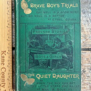 May include: A vintage green book titled "Brave Boy's Trials" with gold lettering and illustrations. The book also features the title "The Quiet Daughter" and the words "Proverb Stories for Boys & Girls". The book is on a wooden surface.