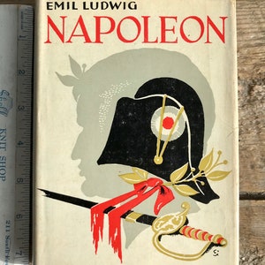 May include: A vintage book cover with a white background and black and red illustrations. The title "NAPOLEON" is in red letters and the author "EMIL LUDWIG" is in black letters. The book is a "MODERN LIBRARY BOOK".