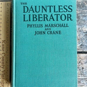 Puede incluir: Un libro de tapa dura titulado "The Dauntless Liberator" de Phyllis Marschall y John Crane. El libro tiene una cubierta de color turquesa con letras negras. Una regla de madera está colocada junto al libro.