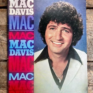 May include: Vintage album cover featuring Mac Davis. The cover has a portrait of a smiling man with dark, curly hair, wearing a white suit with a black collar and a silver necklace. The album title is vertically stacked on the left side.