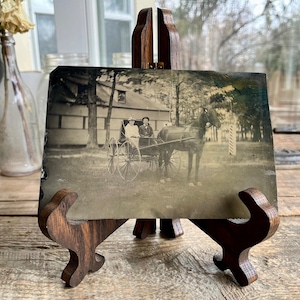 Könnte beinhalten: Ein Schwarzweißfoto einer von einem Pferd gezogenen Kutsche mit zwei Personen darin. Die Kutsche steht vor einem Haus mit Bäumen im Hintergrund. Das Foto ist auf einem Holzständer ausgestellt.