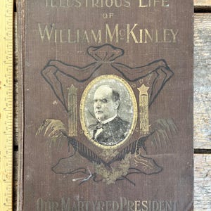 Puede incluir: Un libro marrón con letras doradas titulado "Illustrious Life of William McKinley Our Martyred President". La portada presenta un retrato de William McKinley en un marco ovalado dorado.