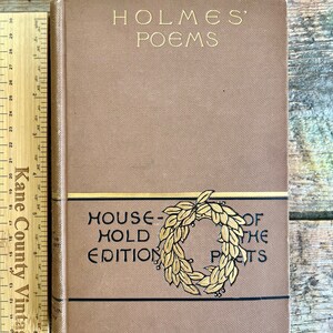 Könnte beinhalten: Ein braunes Hardcover-Buch mit goldenen Lettern auf dem vorderen Umschlag. Der Titel lautet "HOLMES' POEMS" und der Untertitel lautet "HOUSEHOLD EDITION OF THE POETS". Das Buch hat ein goldenes Kranz-Design auf dem vorderen Umschlag.