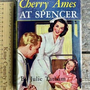 May include: A vintage paperback book cover featuring a nurse, a doctor, and a young girl in a hospital setting. The book title is "Cherry Ames at Spencer" by Julie Tatham.