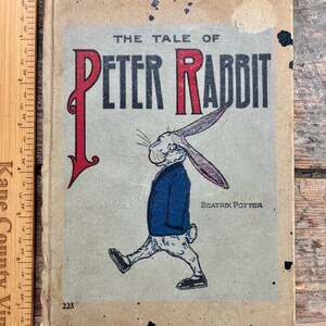 Peut inclure: Ancienne édition de "The Tale of Peter Rabbit" de Beatrix Potter. La couverture présente une illustration de Pierre Lapin en veste bleue. Le titre est en lettres rouges et noires. Le livre est vieilli, avec des signes d'usure visibles.