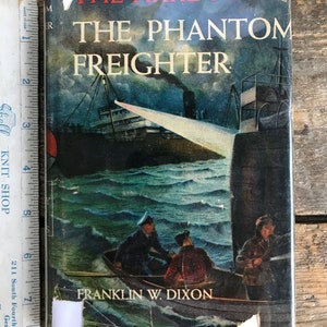 Może przedstawiać: Książka "The Hardy Boys: The Phantom Freighter" z ilustracją morską na okładce. Okładka przedstawia statek, łódź z ludźmi i burzliwe morze. Książka jest owinięta w plastik.