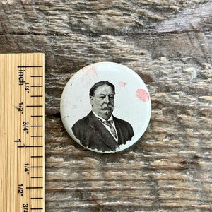 May include: A white button with a black and white image of a man with a mustache and a bow tie. The button has a diameter of approximately 1 inch.