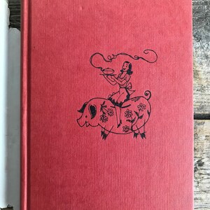 May include: Red hardcover book with a black and white illustration of a woman riding a pig. The woman is holding a plate with a pie on it. The pig is decorated with flowers.