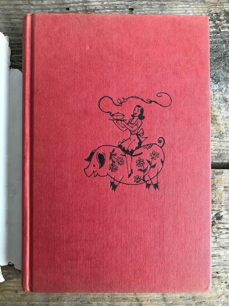 May include: Red hardcover book with a black and white illustration of a woman riding a pig. The woman is holding a plate with a pie on it. The pig is decorated with flowers.