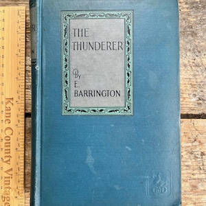 Puede incluir: Un libro antiguo de tapa dura azul titulado "The Thunderer" de E. Barrington. El libro tiene un borde decorativo alrededor del título y el nombre del autor.