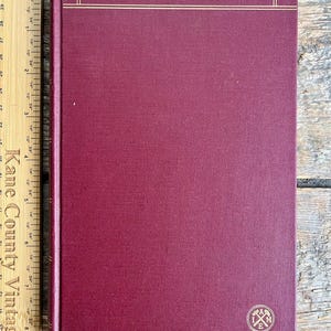 以下が含まれることがあります： 背表紙に金色の文字で「AIME SERIES」と書かれた、えんじ色のハードカバーの本。表紙には金色のエンボス加工のロゴがある。