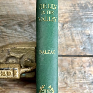 Pode incluir: Um livro antigo de capa dura intitulado "O Lírio do Vale" de Balzac. O livro tem uma capa verde com letras douradas e um emblema decorativo. O livro está posicionado verticalmente contra um fundo de madeira.