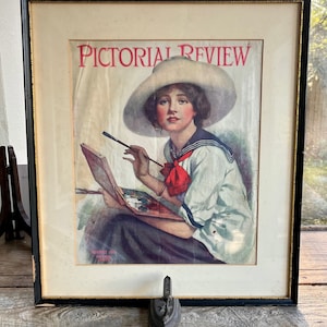 以下が含まれることがあります： 1915年8月号のヴィンテージ雑誌の表紙で、白い帽子とセーラーシャツを着た女性がパレットと筆を使って絵を描いています。雑誌のタイトルは「Pictorial Review」で、50セントです。