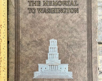 Puede incluir: Una portada de libro vintage con el título "THE MEMORIAL TO WASHINGTON" en letras marrones oscuras. La portada es de color marrón claro texturizado con una ilustración plateada de un monumento. Una regla de madera es visible en el lado izquierdo.