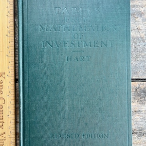 Puede incluir: Un libro antiguo de tapa dura titulado "Tables From Mathematics of Investment" de Hart. El libro tiene una cubierta verde azulada texturizada con letras en relieve. Las palabras "Revised Edition" están impresas en la parte inferior del libro.