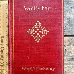 May include: A vintage hardcover book titled "Vanity Fair" by Wm. M. Thackeray. The book has a red cover with gold lettering and a decorative gold emblem. The book is approximately 9 inches tall.